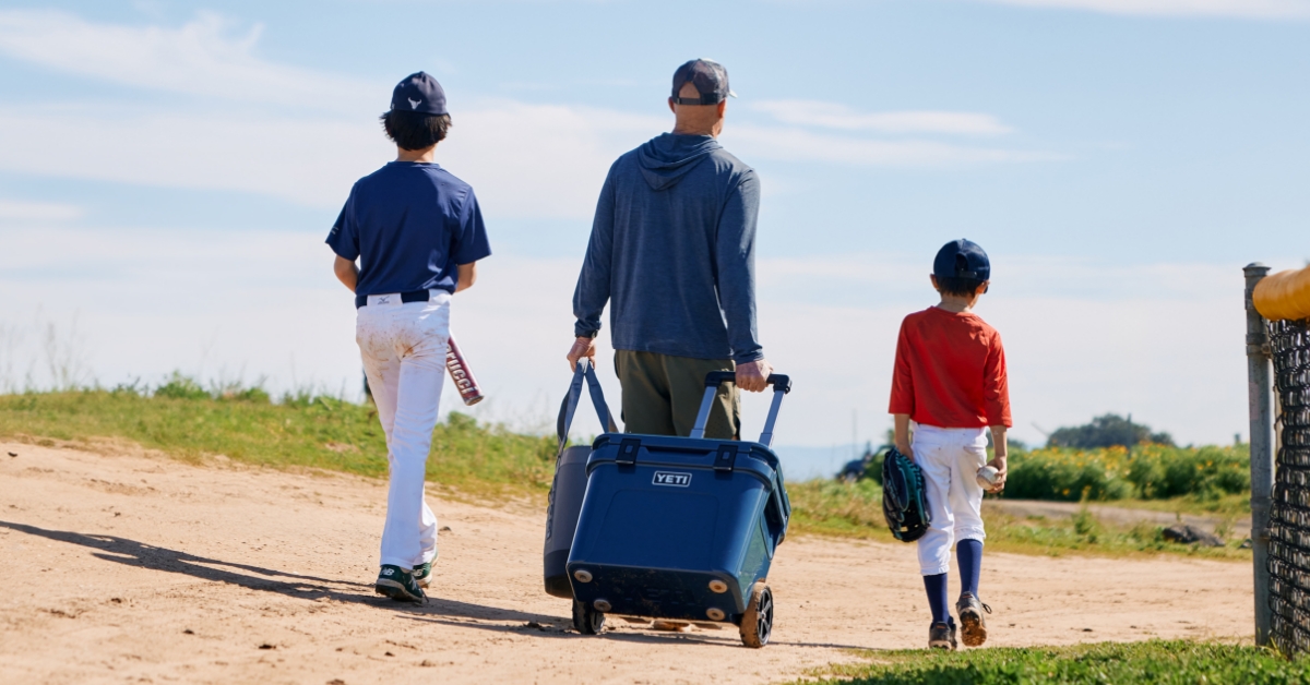 Man wheeling a YETI cooler while two kids in baseball uniforms walk beside him