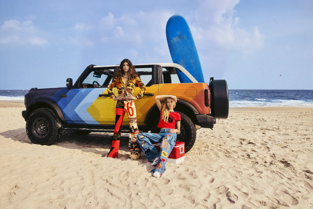 Two women standing beside a colorful Ford Bronco on the beach