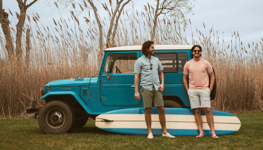 Two men with a surf board next to a vehicle